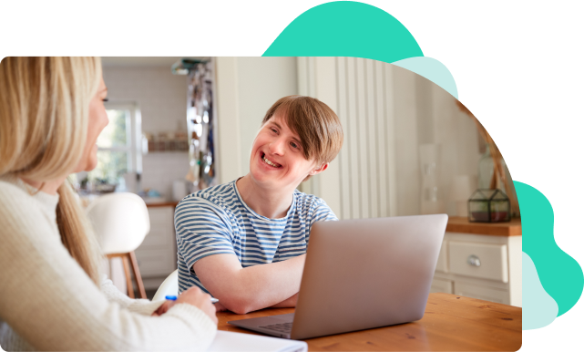 A young male with downs syndrome sitting with a social care worker at a table in a kitchen, using a laptop