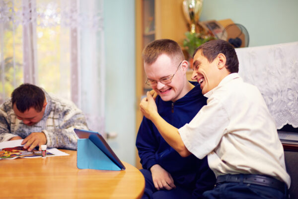 happy friends with disability socializing through internet using a tablet. One male with learning disabilities is in the background reading a magazine