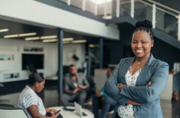 Business woman standing in office with her arms crossed. Behind her are two men and a lady working in the office.