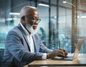 An older black male typing on a laptop, sitting at a desk in an office, surrounded by glass