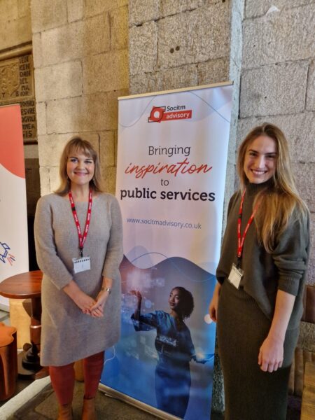 Two female employees stood either side of a pull up banner with the words "bringing inspiration to public services" on it and a photo of a woman working in technology