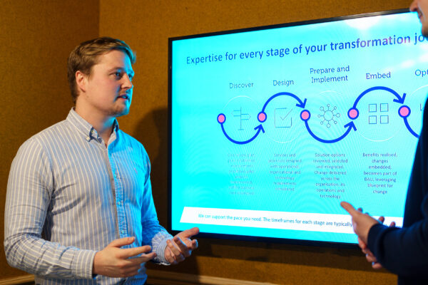 photo of a male colleague standing in front of a screen showing a digital journey diagram, speaking to another colleague off screen to the right