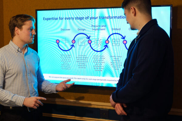 Two male colleagues standing discussing in front of a large television screen showing a six step process
