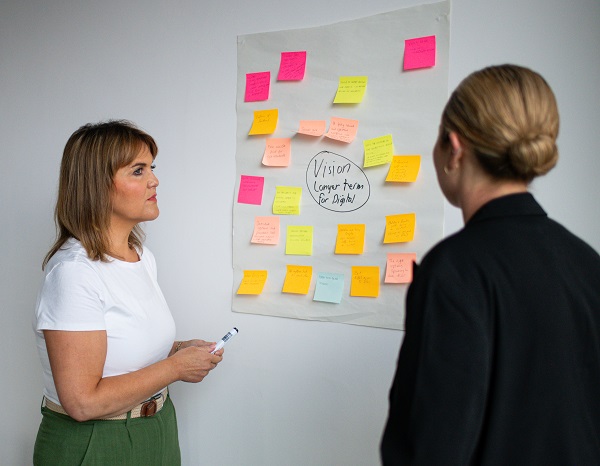 Two women looking at a whiteboard with post it notes on it